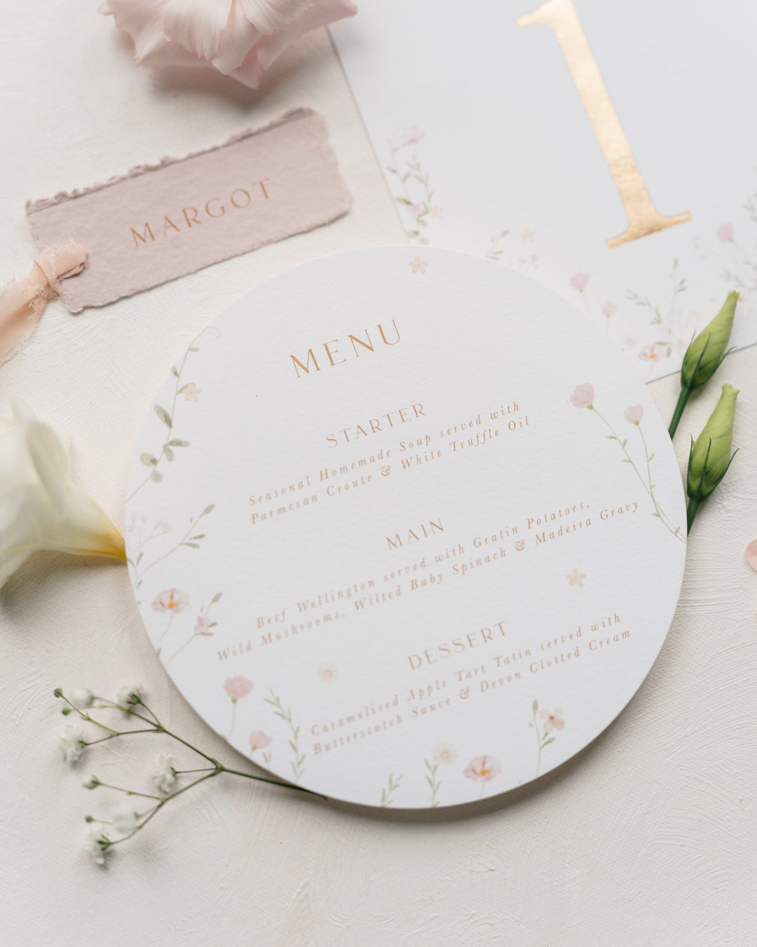 A round wedding menu card with delicate pink floral illustrations and gold text, styled on a white surface with a blush pink name card, pale flowers, green buds, and a gold table number.