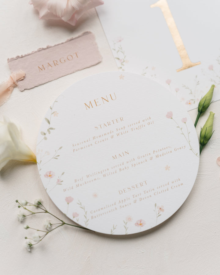 A round wedding menu card with delicate pink floral illustrations and gold text, styled on a white surface with a blush pink name card, pale flowers, green buds, and a gold table number.