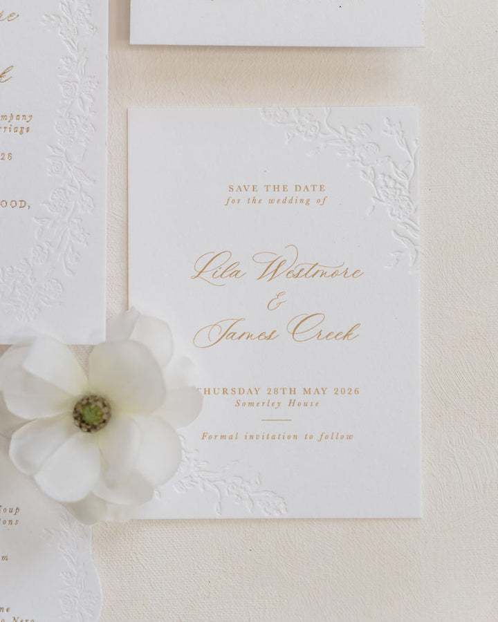 A styled flat-lay of white wedding stationery, including an embossed invitation card, a save-the-date card, a menu card, and a large white flower prop on a textured cream background.