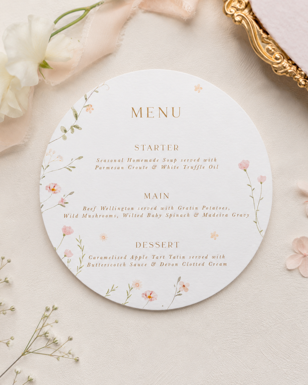 A round printed menu card with delicate floral illustrations and gold text, styled on a light beige surface with soft pink fabric, white flowers, and a gold-framed mirror edge.